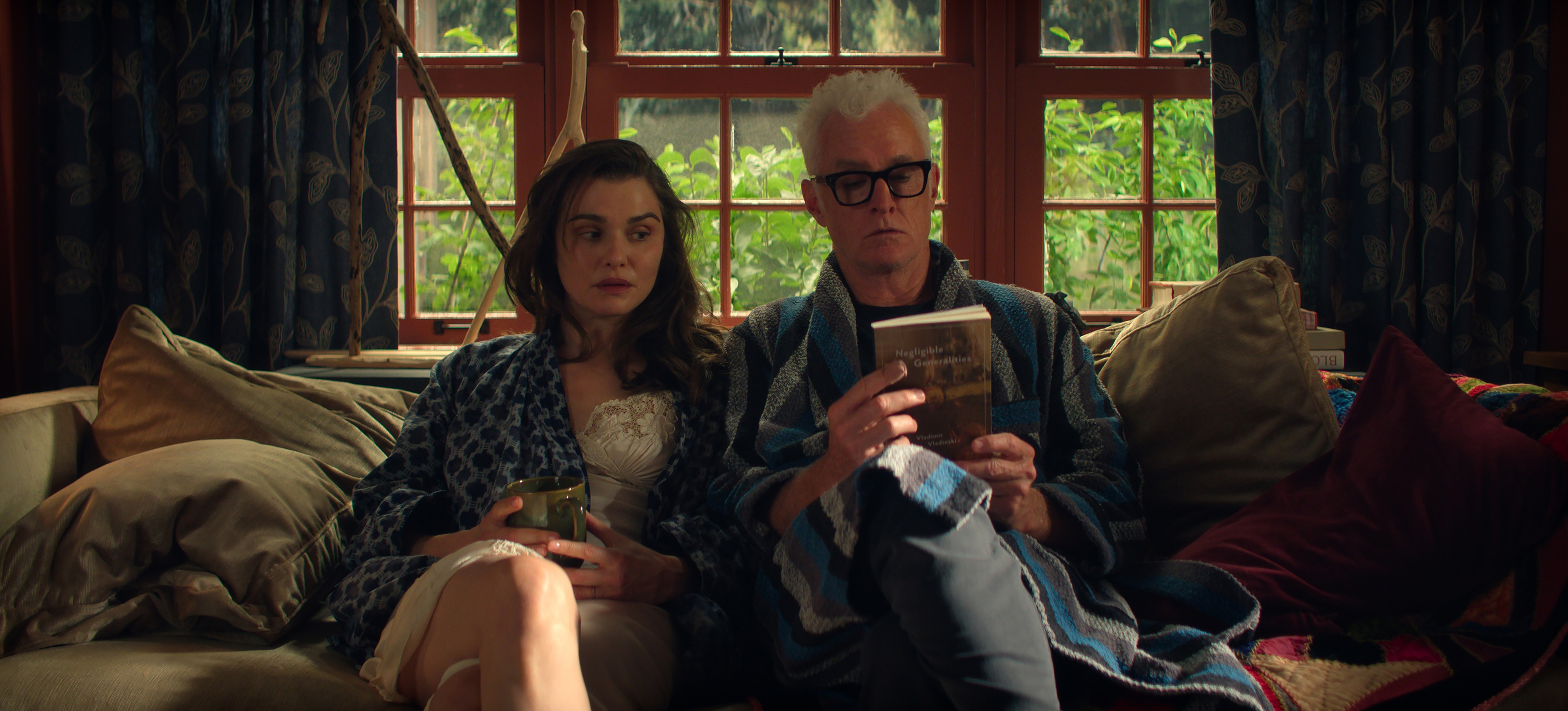 Rachel Weisz as The Protagonist and John Slattery as John sitting on a couch in a cabin in their pajamas in the netflix show vladimir