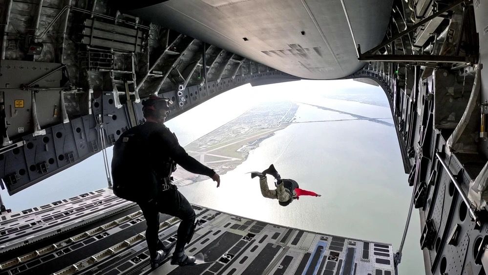 photo from inside a plane, showing one paratrooper jumping out over coastal waters and another preparing to do so