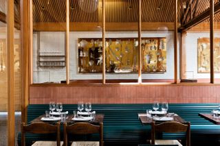 An inventively designed dining room features golden-carved wall art, huge windows, petrol green wooden banquettes, and brasserie-like seating.