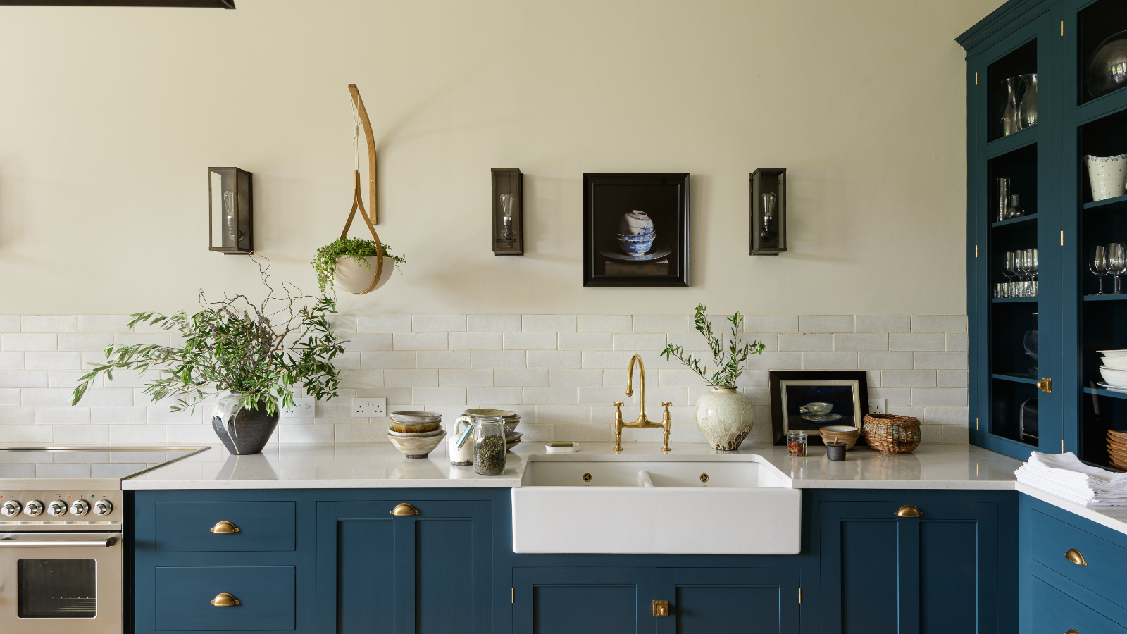 blue and white kitchen, blue cabinetry with white countertops, wall light, counter cabinet with glazing, butler sink, artwork, glass topped stove, brass knobs on cabinets 