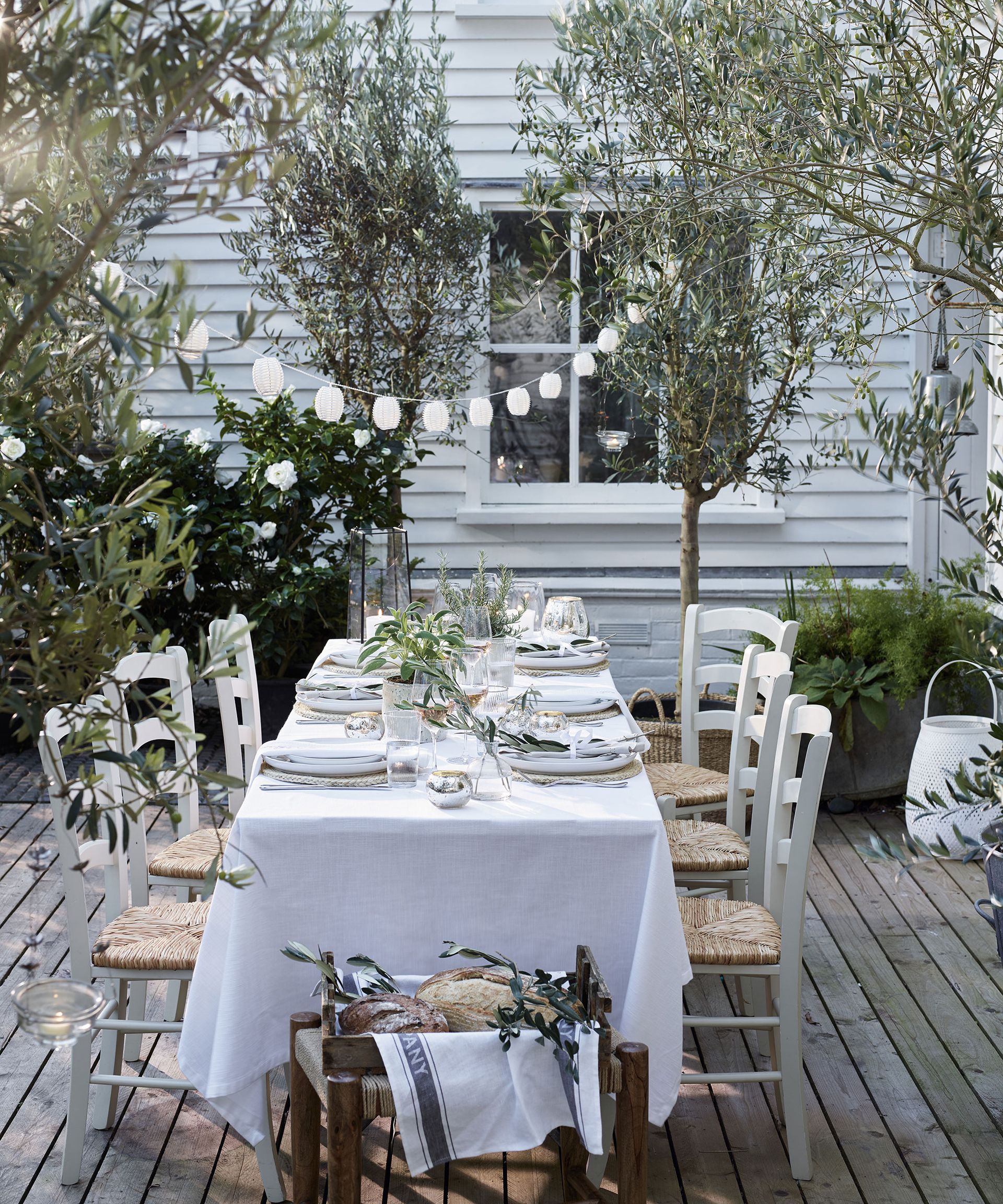 An outdoor dining space with a table covered in a white table cloth and chairs with rattan seats below string lighting
