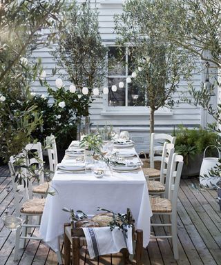 An outdoor dining space with a table covered in a white table cloth and chairs with rattan seats below string lighting