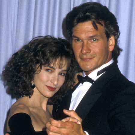 Jennifer Grey wears a black dress while holding a dance pose with Patrick Swayze, her Dirty Dancing co-star who is wearing a tuxedo, as they stand in front of an Oscar statue