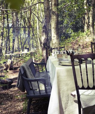 Outdoor dining ideas with dining table in the forest with black dining chairs and grey tablecloth, as well as firepit and serving table