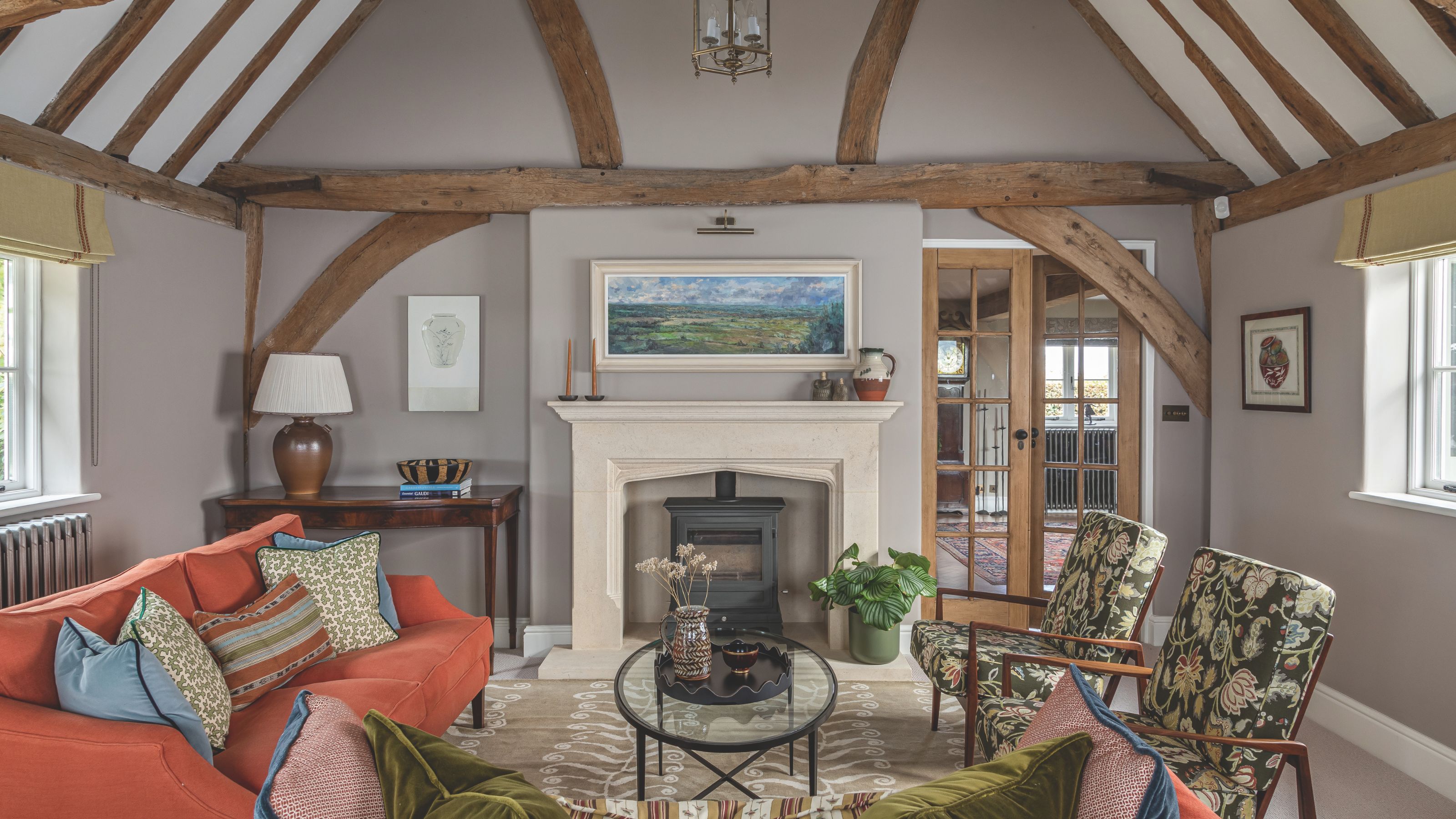 A cozy, rustic living room featuring vaulted ceilings with exposed timber beams and a central stone fireplace. The space is furnished with a burnt-orange sofa, patterned green armchairs, and a round glass coffee table on a neutral patterned rug.