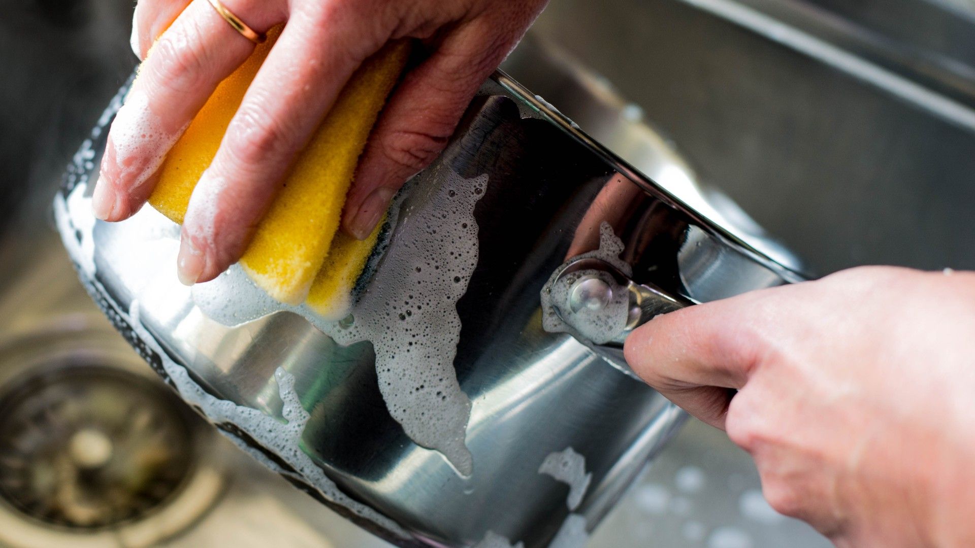 person cleaning a stainless steel pan in hot soapy water with a soft sponge