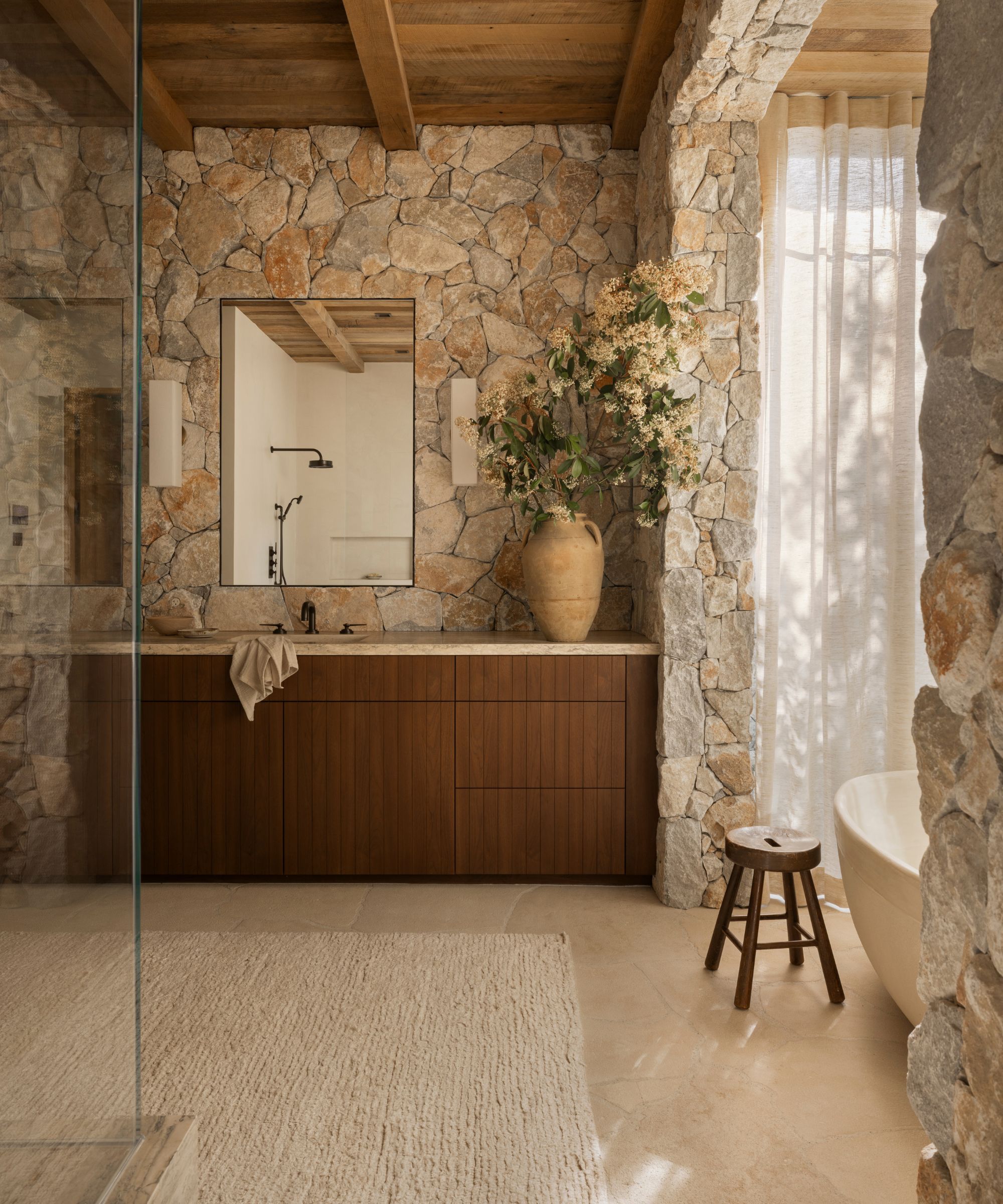 Bathroom with exposed stone walls, wood panelled ceiling, a wood and marble vanity with a sink, a large vase of flowers, and a freestanding bathtub with a wooden stool beside it.