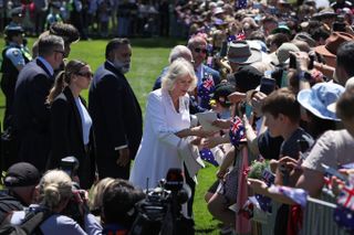 Queen Camilla wearing a white coat shaking hands with a fan in front of a huge crowd of people in Australia