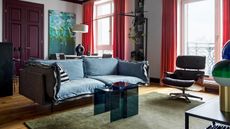 Image of a white living room with hardwood floors and a colorful color palette. There is a green rug, a blue sofa, coral curtains, and a purple door in the room.