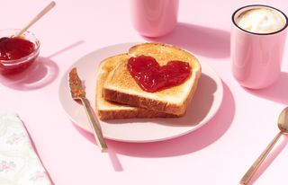 Toast with heart-shaped jam on, on a pink plate with a pink mug of coffee, on a matching pink background.