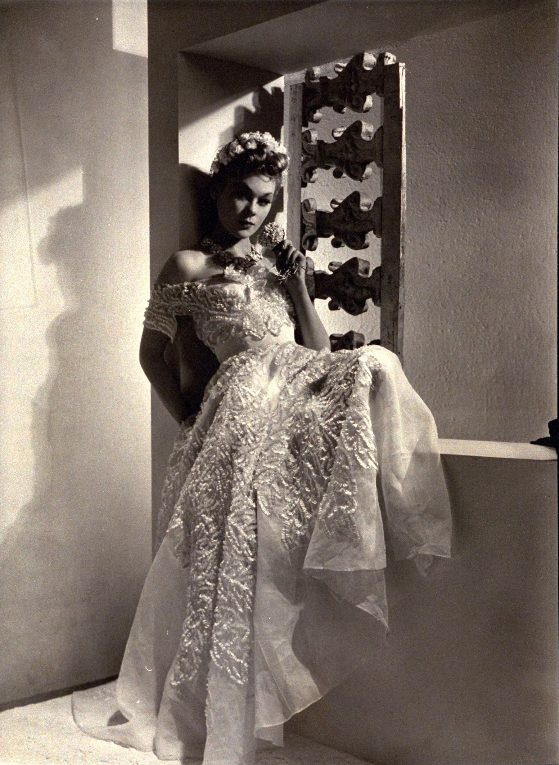 Model wearing a floor-length evening gown of white organdie, decorated all over the full skirt and off-the-shoulders bodice with white ribbons, by Chanel; a necklace of white china camellias; and floral headband