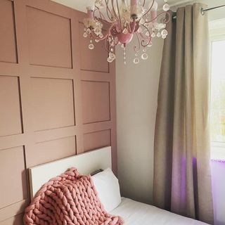 a young girls room with a pink panel wall, pink chandelier above a single bed and brown full length curtains in front of the window