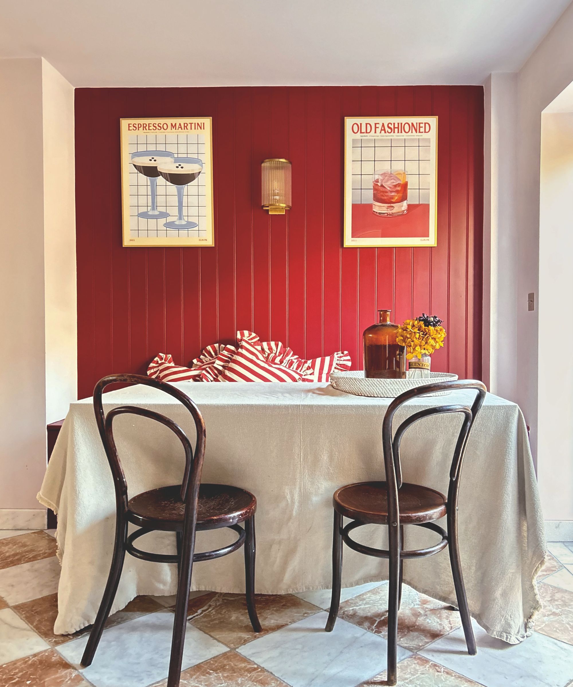 A dining area with a red shiplap wall featuring "Espresso Martini" and "Old Fashioned" art, a linen-covered table, bentwood chairs, and a checkered marble floor.