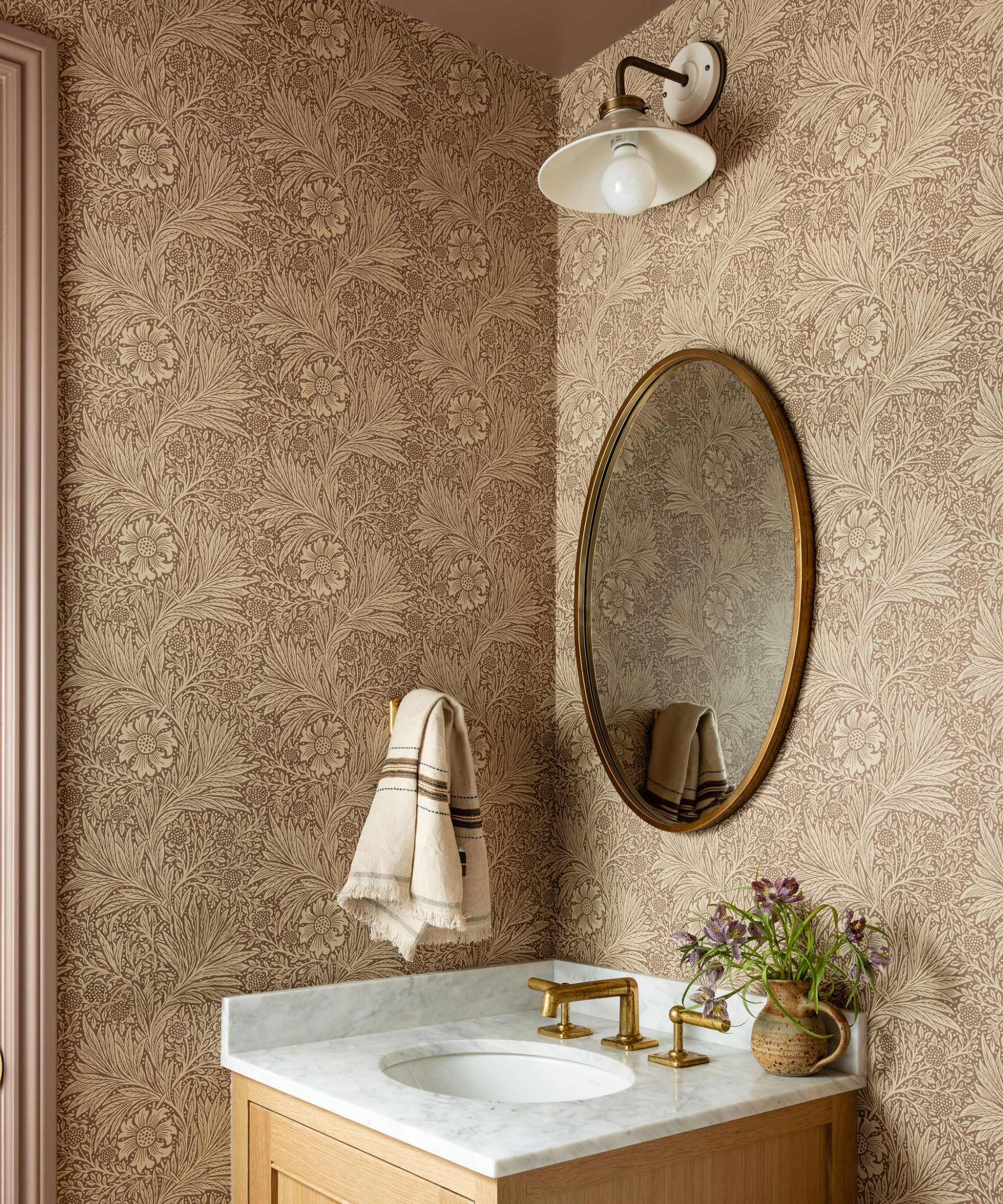 Guest bathroom with floral wallpaper, an oak vanity, marble countertop, brass faucet, oval mirror and cream wall light.