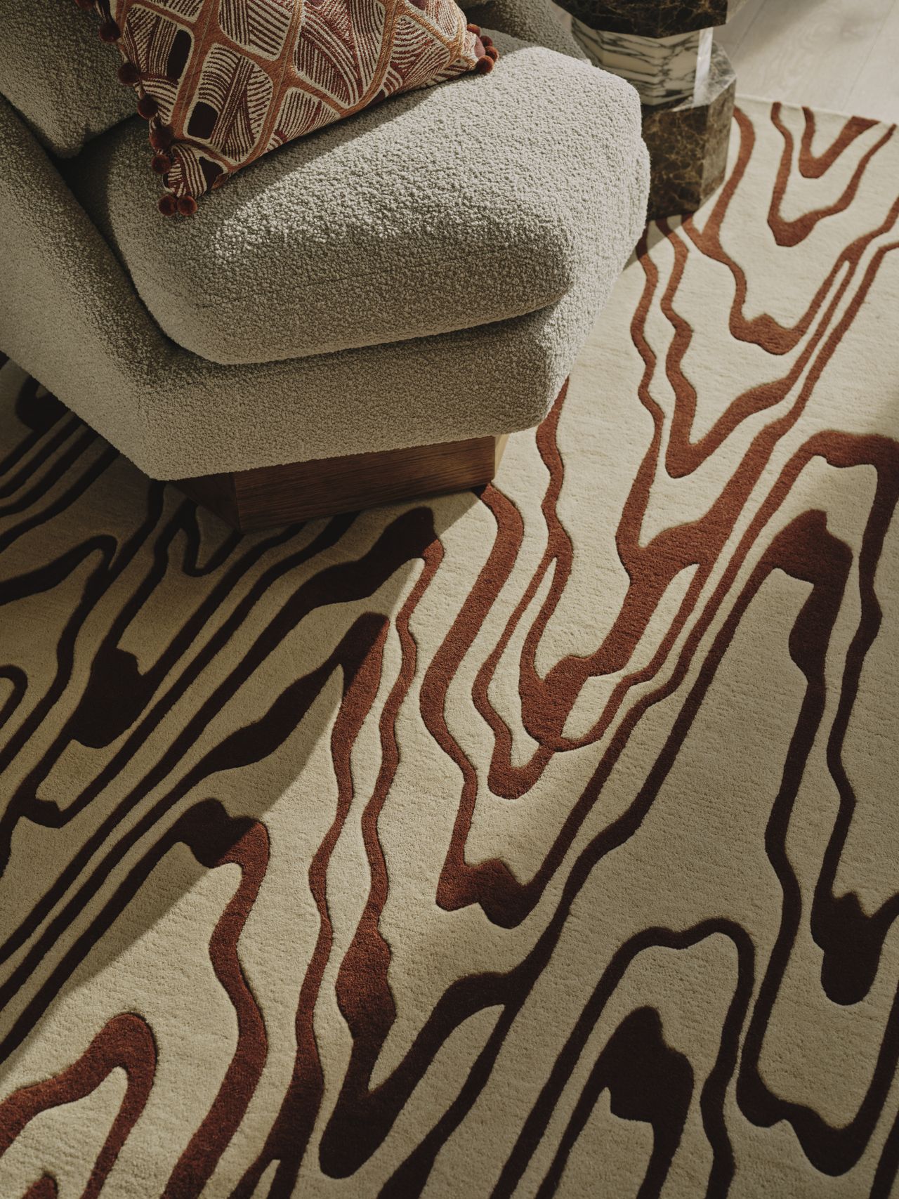 Image from above of a white and terracotta rug in a wavy line pattern. The seat of a boucle, off-white accent chair is in view with a patterned pillow resting on the seat.