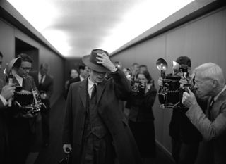 cillian murphy as robert oppenheimer walking through a long hall with people taking pictures in the movie oppenheimer