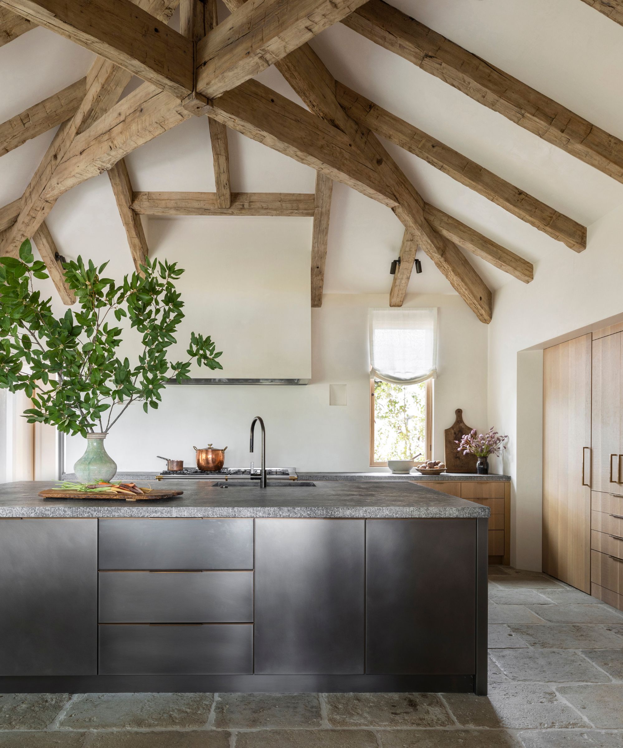 A large kitchen with tall ceilings featuring exposed wood beams, white walls, no backsplash, wooden cabinets, and a stainless steel island