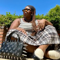 chichi wears black and white tank top and plaid skirt with black and white loafers and spiked bag while sitting on the ground 