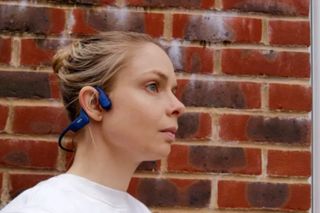 This shows a pair of headphones you could use when cycling. The Shokz OpenRun headphones are bone conducting and are shown on a woman wearing a white jumper standing in front of a brick wall.