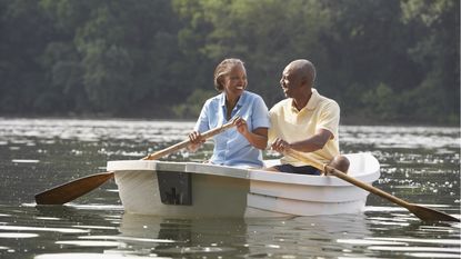 An older couple laugh while paddling a small boat on a lake.