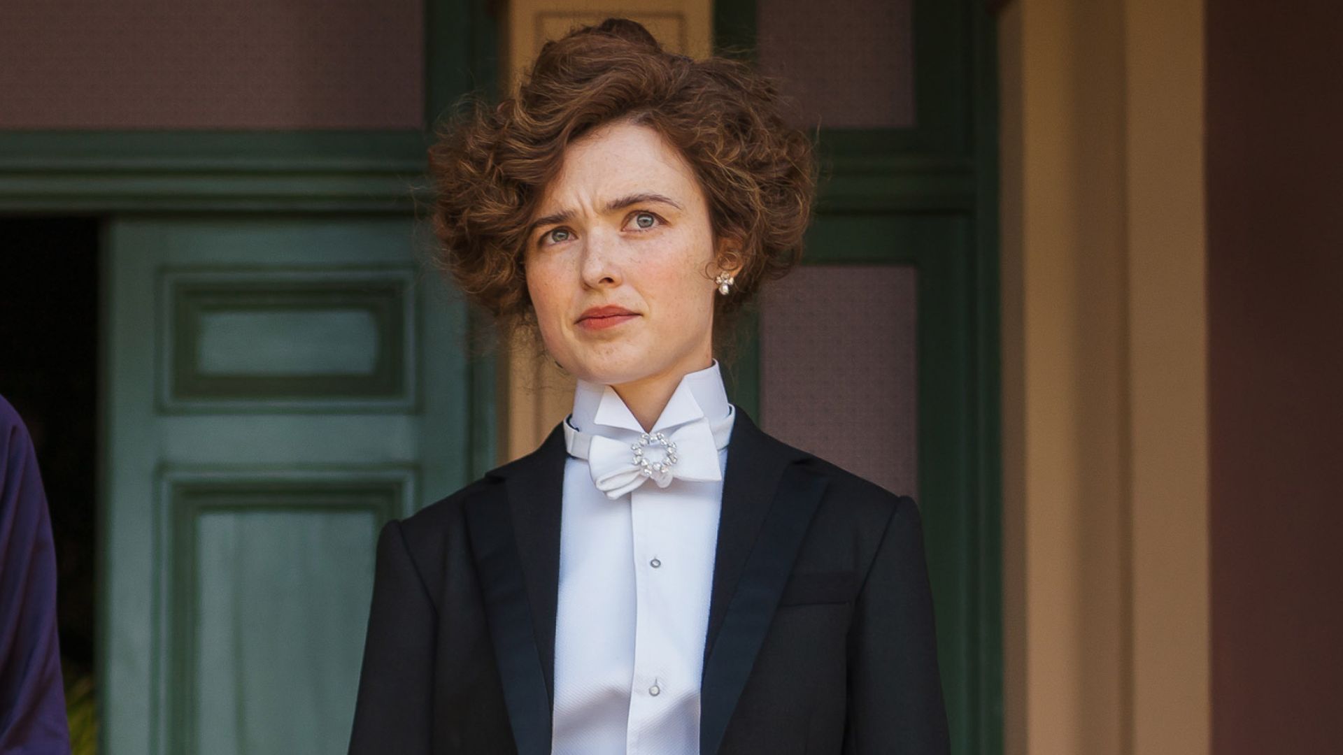 Sybilla wears a suit, standing in a doorway