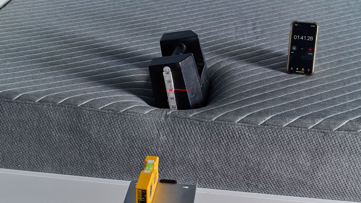 A close up of a 25kg weight balanced on the side of the Siena Mattress in our sleep studio. A tape measure is attached to the side of the weight. In the front of the shot is a laser measure, balanced on a tripod. The laser is aimed at the tape measure with the line indicating how far the weight sinks into the mattress. To the right, a phone is displaying a timer indicating the test has been taking place for 1 minute 41 seconds.