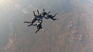 five skydivers hold each others' arms high above a desert landscape.
