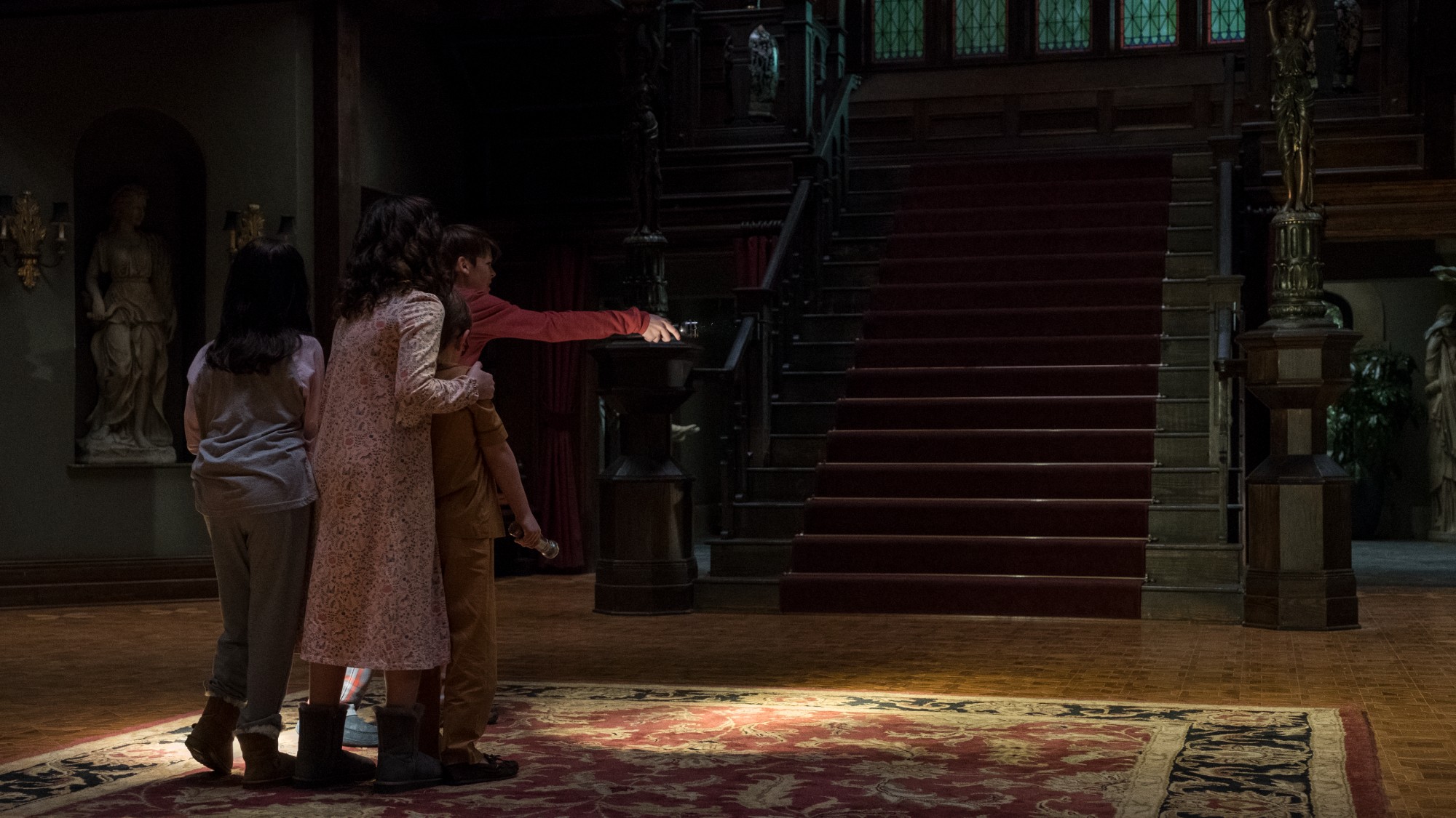 a rear shot of a group of four children in their pajamas in front of an enormous wooden staircase in an old house. one of the children points a flashlight to the right