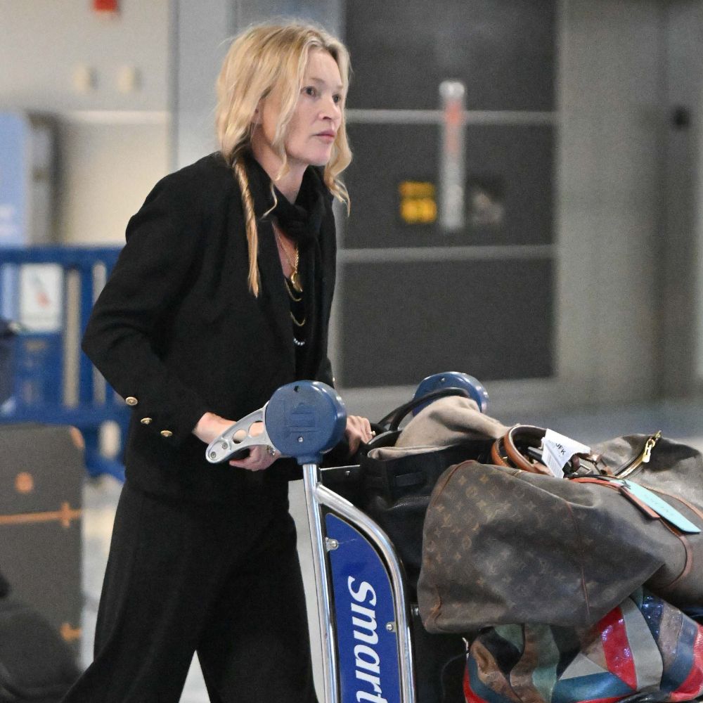 Kate Moss pushes a luggage trolley through the airport wearing black trousers with a black blouse.