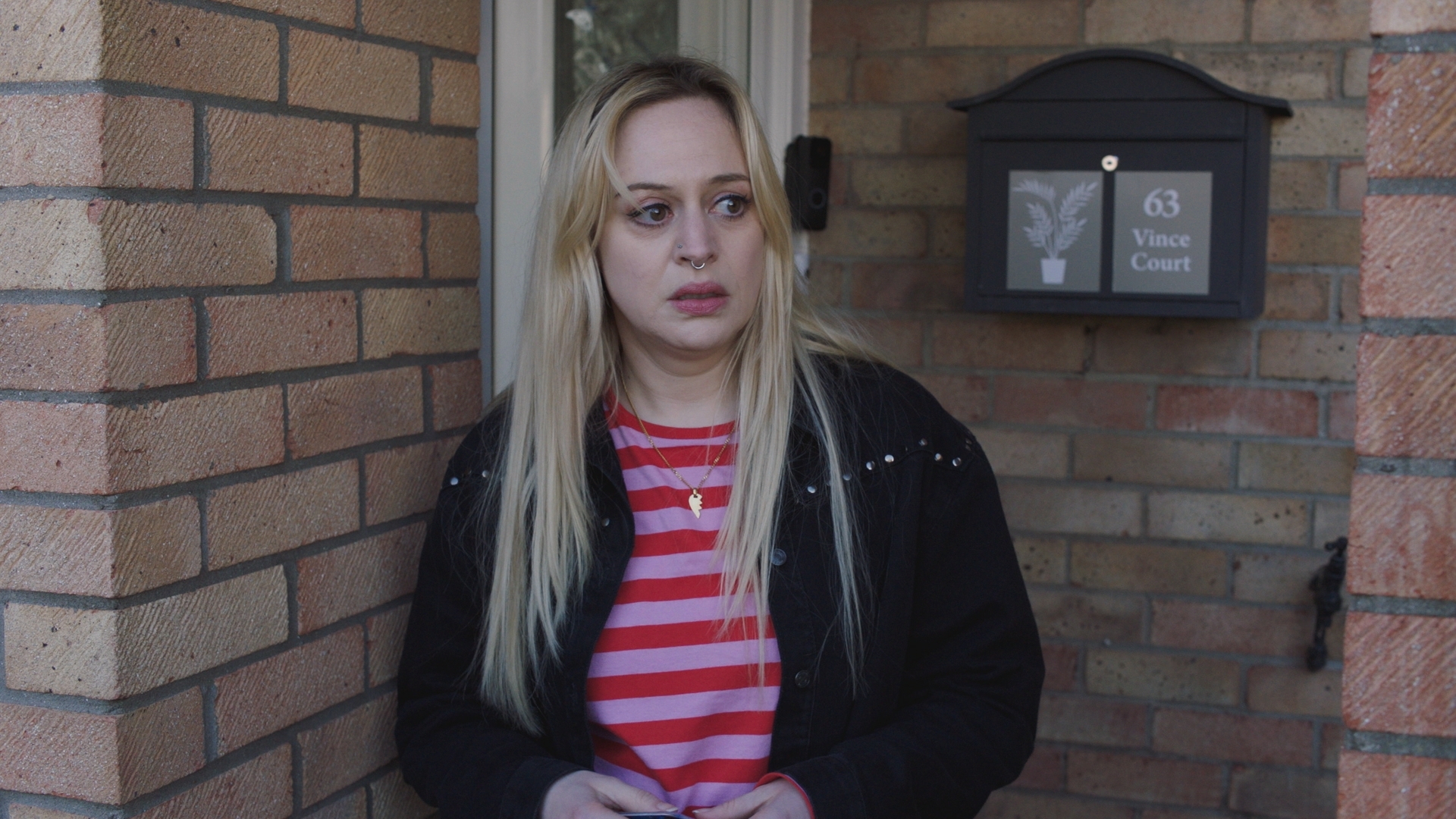 Nicole Piper wearing a black jacket and pink stripy top standing outside her mother's front door looking scared.