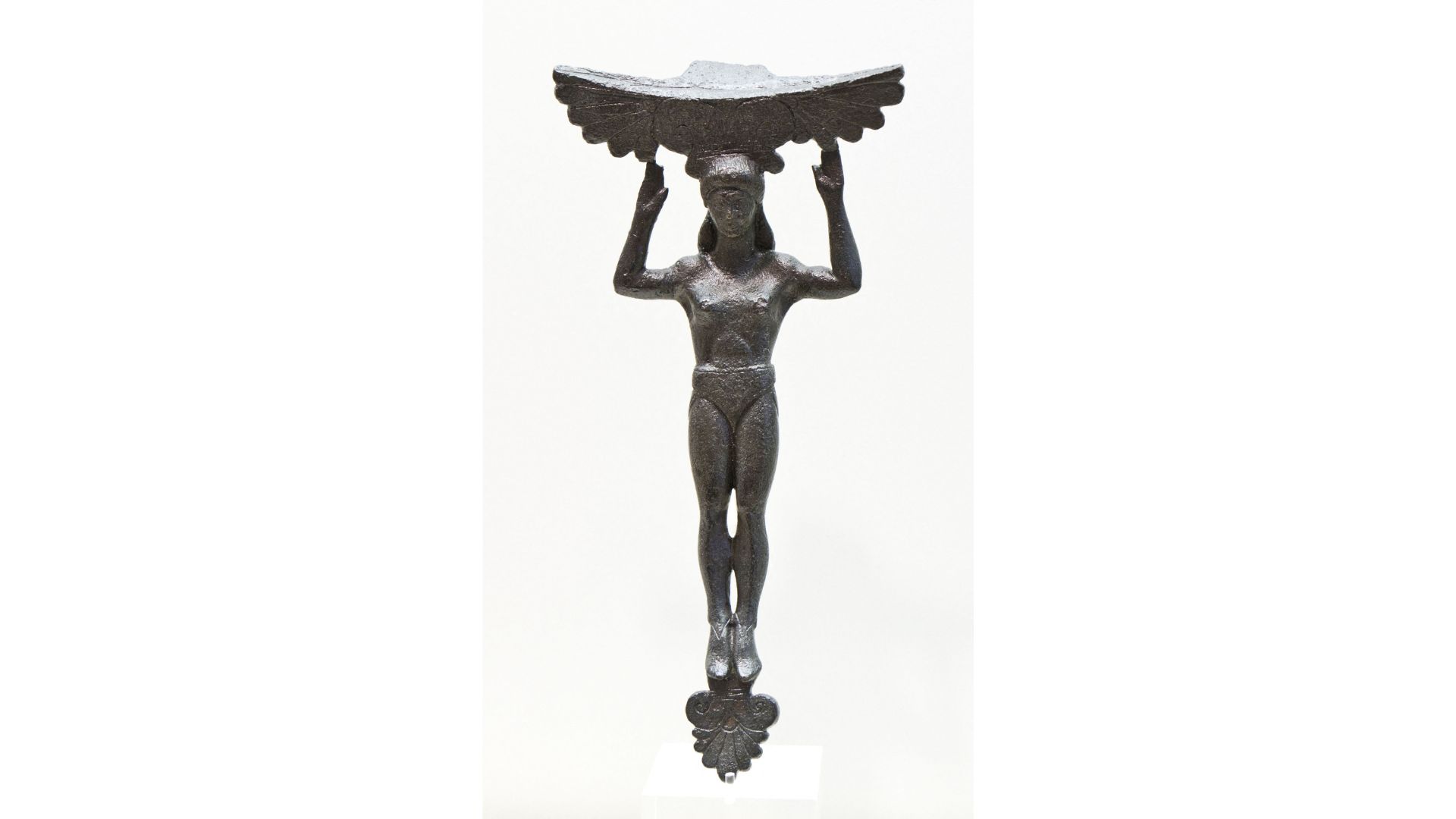 Photo of a bronze statuette of a woman standing upright with her legs together and arms bent at the elbows and pointing up. Her fingertips are touching a feathered decoration that sits above her head.
