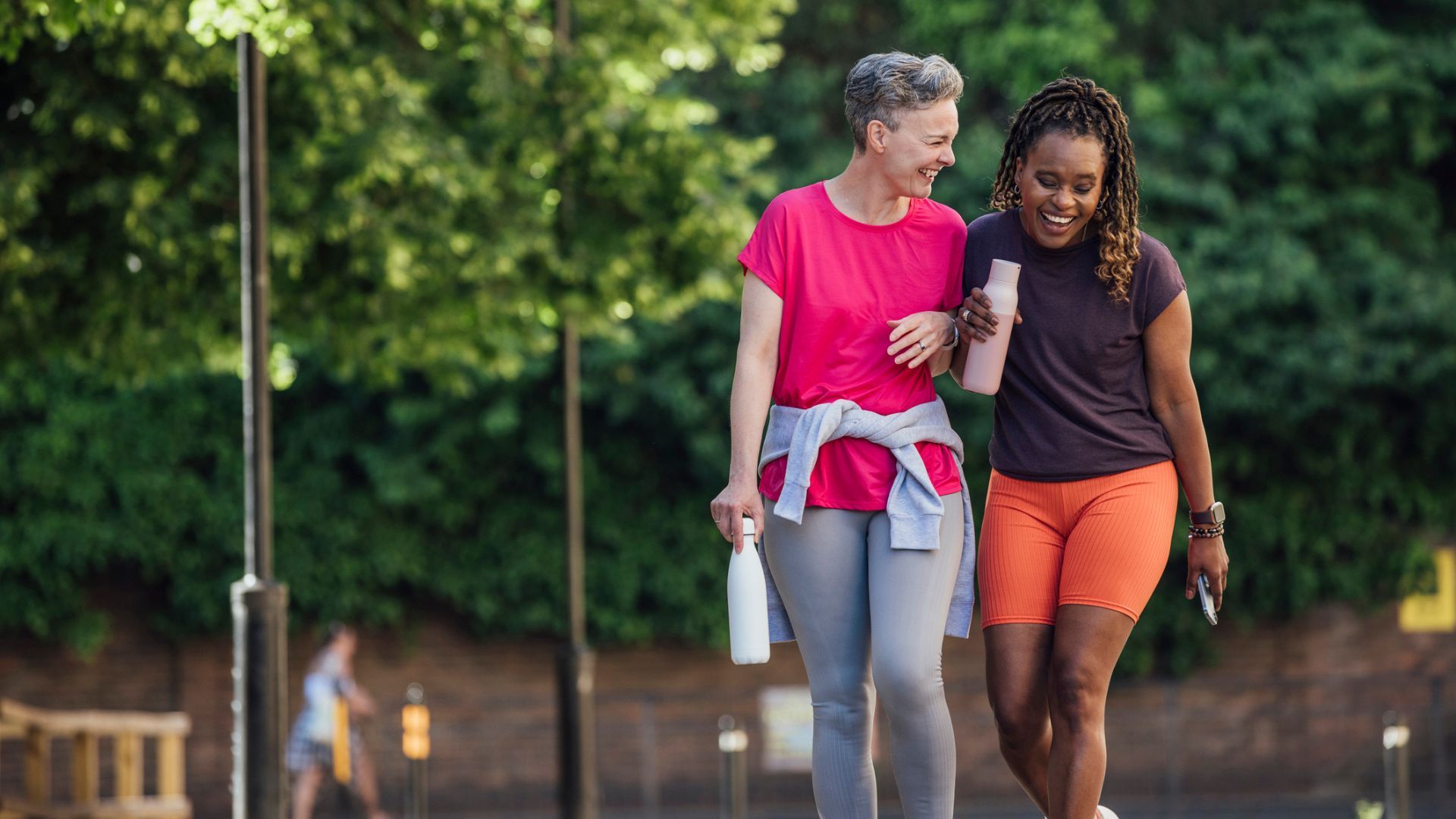 Two women laughing and walking together in workout clothes and holding water bottles