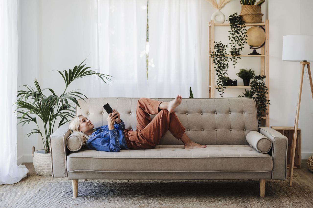 A woman using a phone while lying on a cream sofa surrounded by house plants and a side lamp.