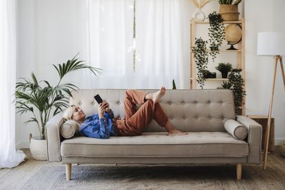 A woman using a phone while lying on a cream sofa surrounded by house plants and a side lamp.