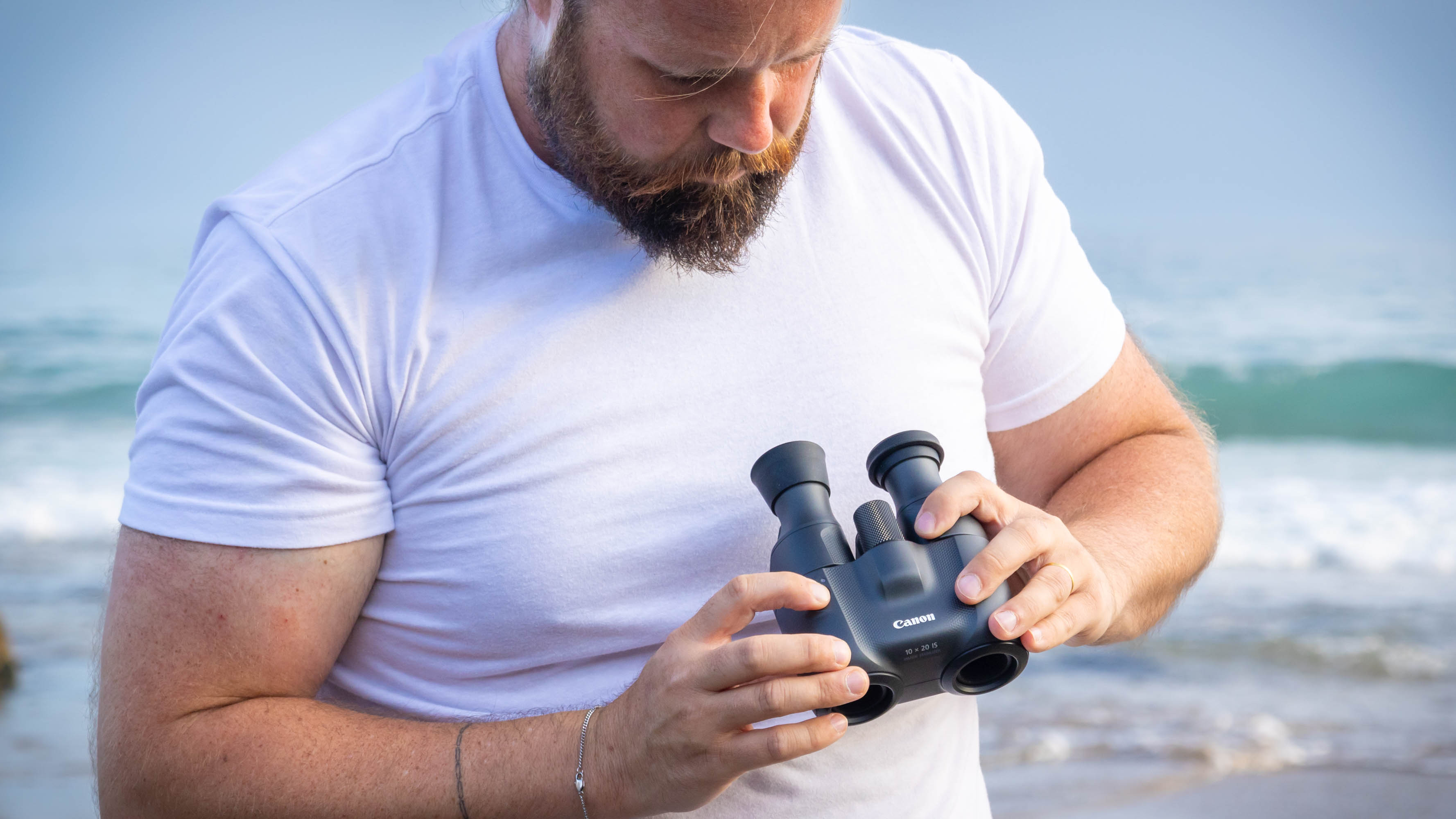 A male holding and looking at the Canon 10x20 IS binoculars with the sea behind them.