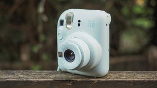 An Instax Mini 12 camera, on a wooden surface in a woodland setting