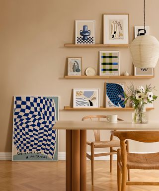 A light brown dining room with three wooden shelves with wall art prints on them, a light wooden dining set with a rectangular table and two chairs, a cream pendant lamp, and a blue and white checked print on the floor