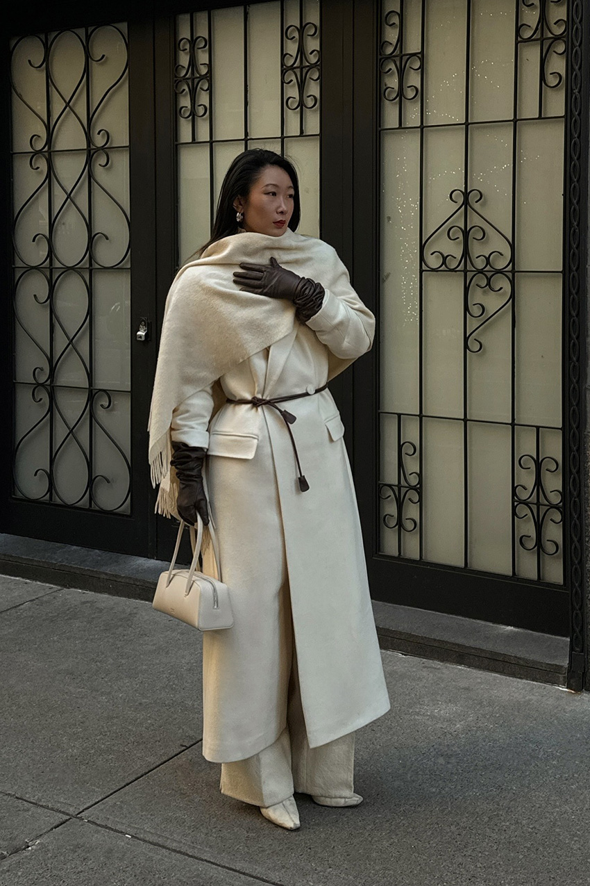 A woman standing on the sidewalk wearing a long white coat styled with a long white scarf, a brown woven belt, brown opera leather gloves, ivory wide-leg trousers, white pointed ankle boots, silver hoops, and a white barrel bag.