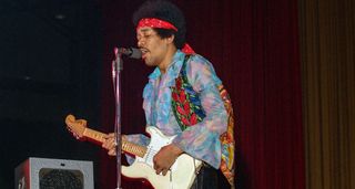 Jimi Hendrix wears a red bandana and a patterned shirt as he plays his Fender Strat live in 1970.