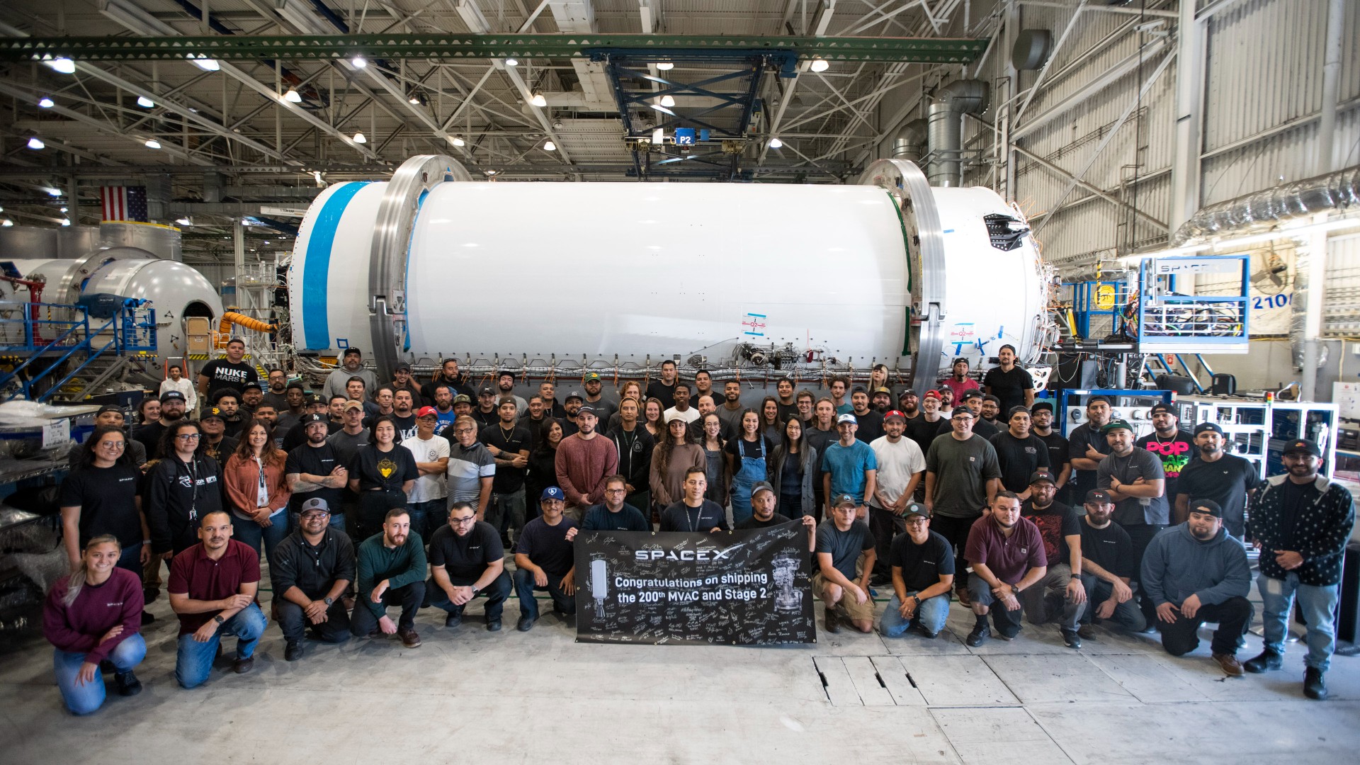 dozens of people smile for a portrait in front of a large white cylinder in a massive warehouse