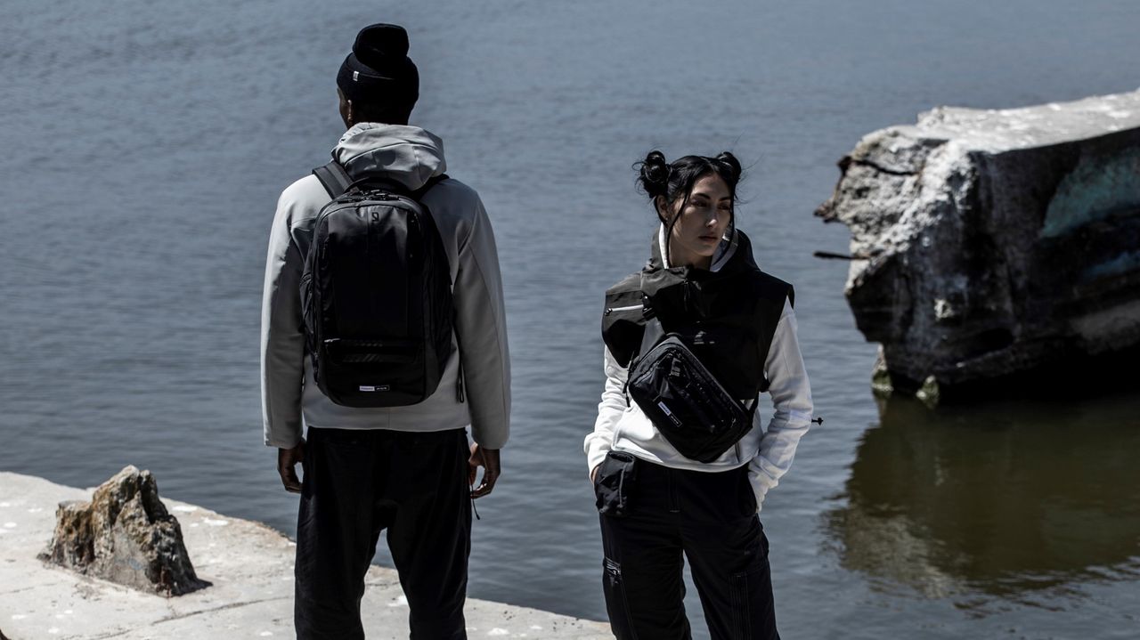 Two models. Female facing front with cross body bag and male model facing out to the ocean with back back.