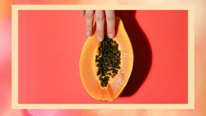 Female hand and half of fresh papaya with black seeds on red background in a summer day. Erotic concept. Top view with copy space.