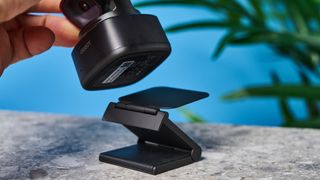 a small PTZ webcam with 4K resolution and a magnetic attachment clip is photographed in front of a blue background and a green plant