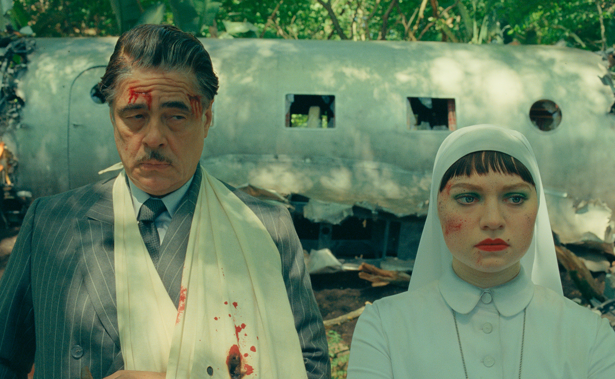 Still from the film The Phoenician Scheme of Benicio del Toro and Mia Threapleton. Both are in character standing in front of damaged plane. They have scrapes and blood on their faces.