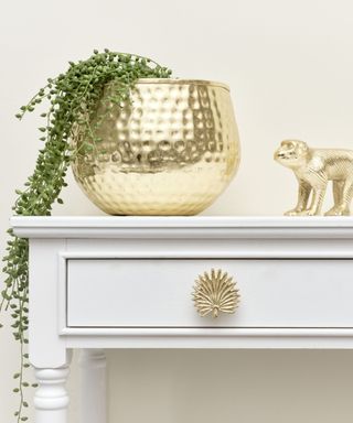 A white drawer with a gold knob, with a gold plant pot and monkey decoration on top of it