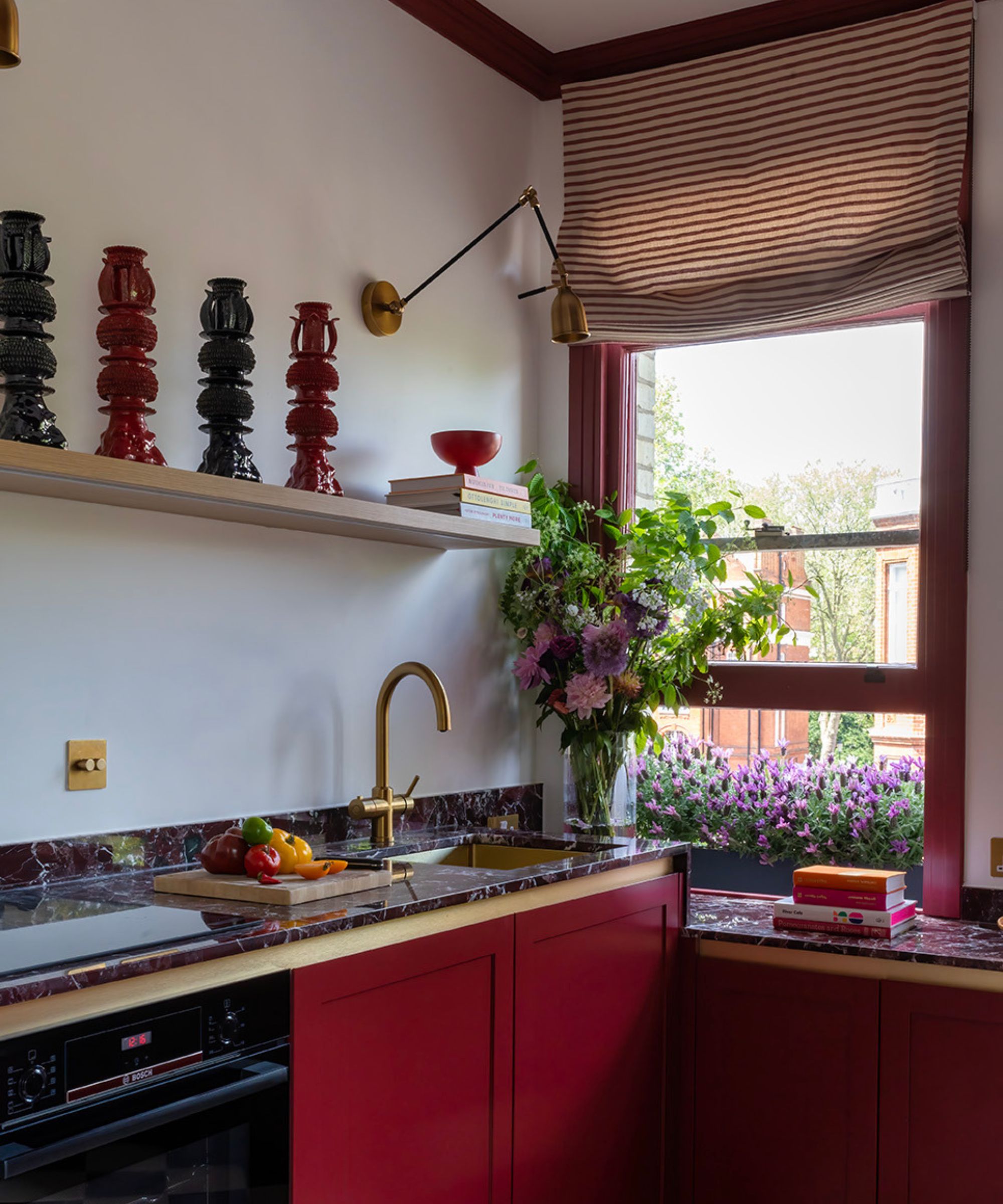 A kitchen with white walls, dark red cabinets, Merlot-toned countertops, window frames, and decor, with wood and brass details