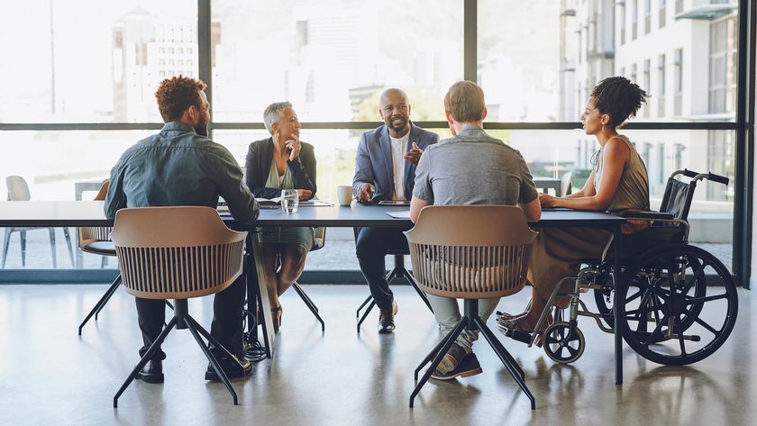 A diverse team sat at a boardroom table, with a female worker in a wheelchair positioned at the head of the table to the right of frame.