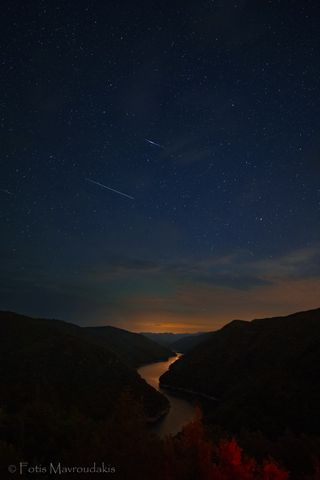 perseid meteors shoot across the sky above a river and mountains.