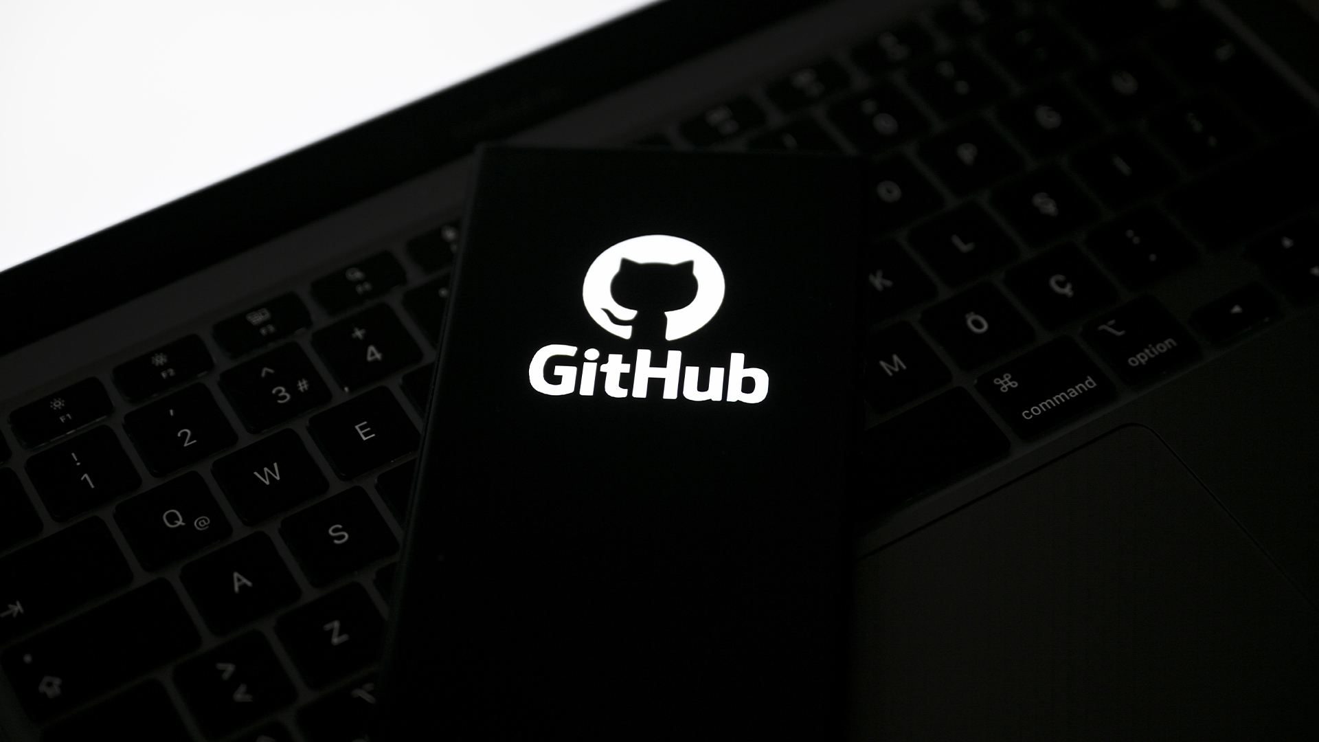 GitHub logo pictured on a smartphone placed on top of a laptop keyboard, with blank white screen in corner.