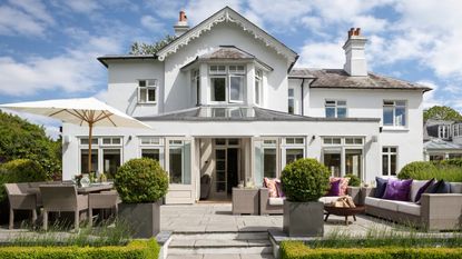 A white house exterior with a large paved patio, with a seating area under a parasol, wicker sofa with cushions and firepit. There are large plants in grey square pots, and central steps in the foreground. Overhead are blue, cloudy skies.